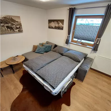 Ein gemütliches Zimmer mit einem Schlafsofa und einem Fenster. Die Wände sind mit Bildern dekoriert und der Boden ist aus Holz.