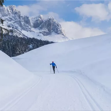 Ein Langläufer auf einem schneebedeckten Weg in den Bergen. Die Landschaft ist ruhig mit schneebedeckten Hügeln und klaren Himmel.