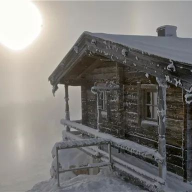 Eine verschneite Holzblockhütte steht in einer nebligen Landschaft. Die Sonne scheint durch den Nebel und erzeugt eine mystische Atmosphäre.