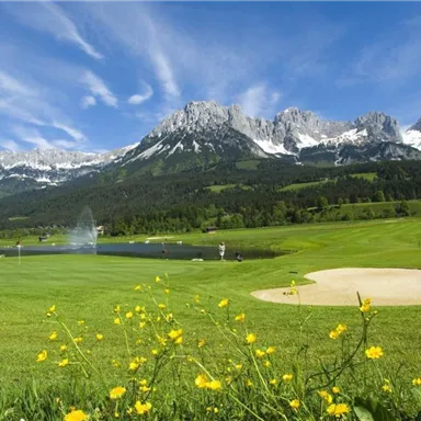 Eine malerische Golfanlage mit grünen Wiesen und blühenden Blumen. Im Hintergrund sind majestätische Berge und ein See mit einem Wasserspeier zu sehen.