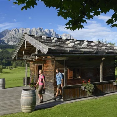Eine wunderschöne Berghütte umgeben von grünen Wiesen und majestätischen Bergen. Zwei Personen spazieren entspannt zur Hütte.