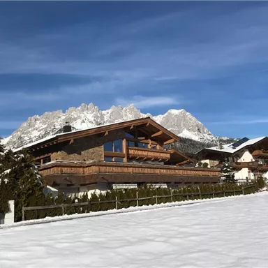 A picturesque winter landscape with snow-covered hills and traditional chalets. In the background, impressive mountains can be seen under a clear blue sky.