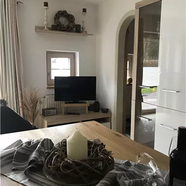 Ein gemütlicher Wohnraum mit einem Holztisch, einem Fernseher und einer modernen Wohnwand. Die Dekoration umfasst eine Kerze und eine stilvolle Tischdecke.