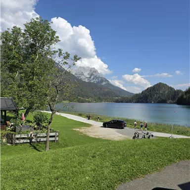 A quiet lake surrounded by mountains and trees. The meadow is green and some people can be seen at the shore.