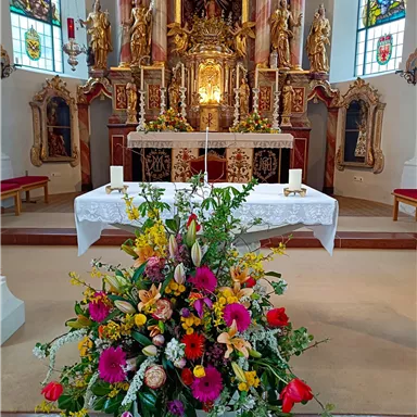 A decorated altar in a church with a large flower arrangement in the foreground. The walls are decorated with colorful stained glass motifs.