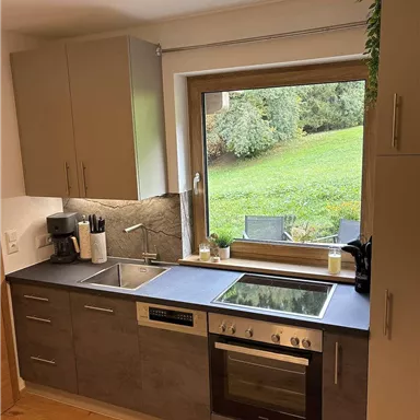 Eine moderne Küche mit einer großen Fensterfront, die einen Blick auf den Garten bietet. Der Raum ist hell und gut ausgestattet mit Kochgeräten.