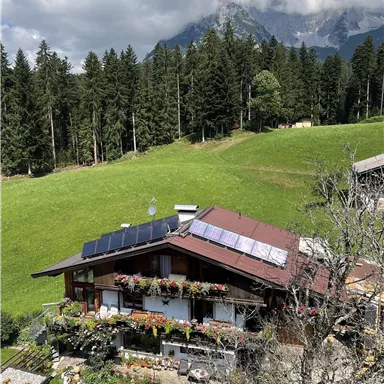 A charming house surrounded by green meadows and tall trees. In the background, majestic mountains rise under a partly cloudy sky.