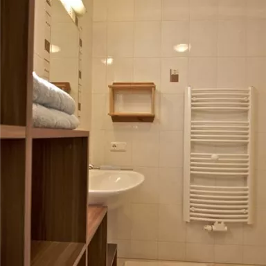 A modern bathroom with bright tiles and a sink. On the wall, there is a towel warmer and a shelf with folded towels.