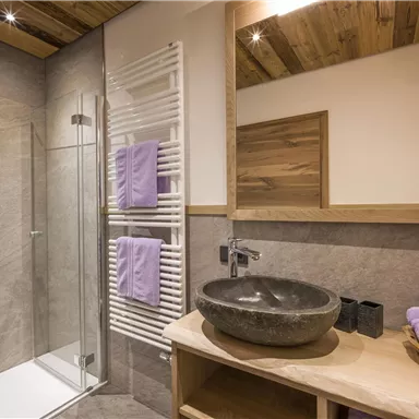 Ein modernes Badezimmer mit einer Glasdusche und einem Waschbecken aus Stein. Handtücher in lila und Holzdetails verleihen dem Raum eine gemütliche Atmosphäre.