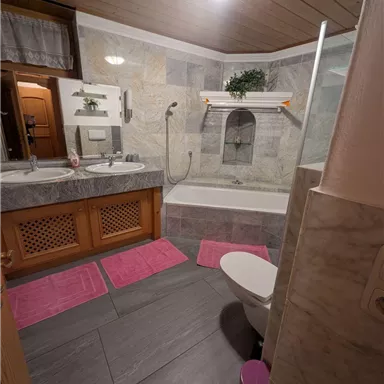 Ein modernes Badezimmer mit einer Badewanne und Dusche. Die Wände sind mit Steinfliesen verkleidet, und pinkfarbene Teppiche sorgen für einen farbenfrohen Akzent.
