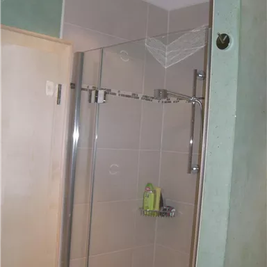 A modern shower with a glass partition and bright tiled floor. On the wall are toiletries and a rain shower above.