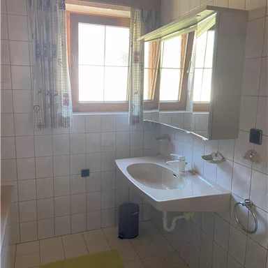 Ein modernes Badezimmer mit einem Waschbecken, einem Spiegel und zwei Fenstern. Der Boden ist gefliest und ein grüner Badeteppich liegt vor dem Waschbecken.