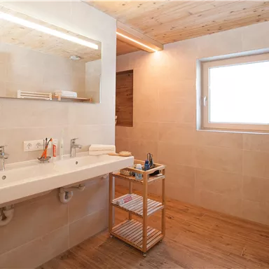 Ein modernes Badezimmer mit hellem Holz und elegantem Fliesenboden. Die große Spiegelleiste und Fenster sorgen für viel Licht.