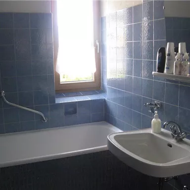 A bathroom with blue tiles, a bathtub, and a sink. The window lets in plenty of daylight.