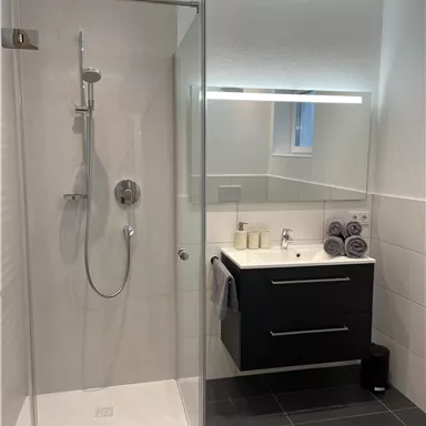 A modern bathroom with a glass shower and an elegant sink. The walls are light and the floor is dark tiles.