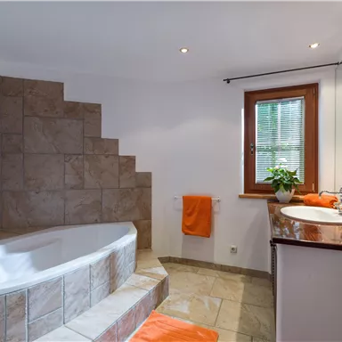 Ein modernes Badezimmer mit einer komfortablen Badewanne und einem stilvollen Waschbecken. Orangefarbene Handtücher und Pflanzen sorgen für einen einladenden Akzent.