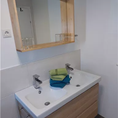 A modern bathroom with a double sink and a wooden mirror. Colorful towels are neatly stacked on the sink.