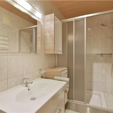 A modern bathroom with a shower cabin and a sink. The walls are light tiled and there are large mirrors.