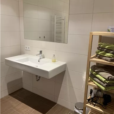 A modern bathroom with a sink and a large mirror. Next to the sink is a shelf with clean towels.