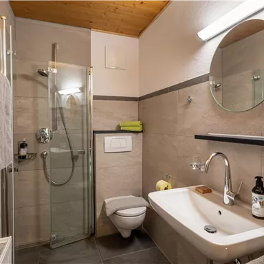 Ein modernes Badezimmer mit einer Glasdusche und einem Rundspiegel. Es sind Handtücher, Pflegeprodukte und ein Fön sichtbar.