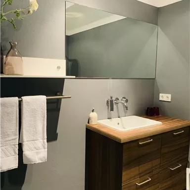 Ein modernes Badezimmer mit einem schlichten Waschbecken und einem Holzunterbau. Weiße Handtücher hängen neben einem Spiegel an der Wand.