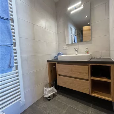 A modern bathroom with a wooden dresser and a mirror. Towels are hanging on the wall and the floor is made of dark tiles.