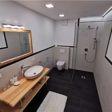 A modern bathroom with an elegant shower and a large mirror. The floor is covered with dark tiles and a wooden shelf is decorated with toiletries.