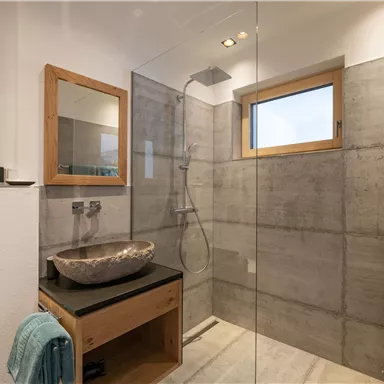 A modern bathroom with a spacious shower and an elegant sink. The walls are made of concrete, and there is plenty of natural light coming through the window.