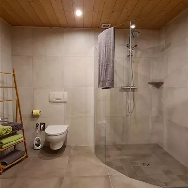Ein modernes Badezimmer mit einer großzügigen Dusche und einem WC. Die Wände sind in neutralen Farben gehalten und es gibt frische Handtücher und einen Handtuchhalter aus Holz.