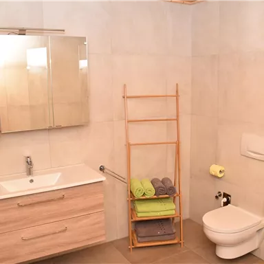 Ein modernes Badezimmer mit einem Waschbecken, einem Spiegel und einem WC. Handtücher sind auf einem Holzregal ordentlich gestapelt.