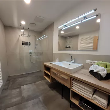 A modern bathroom with a large shower and a sink on a wooden unit. The walls are in neutral colors and the lighting creates a bright atmosphere.
