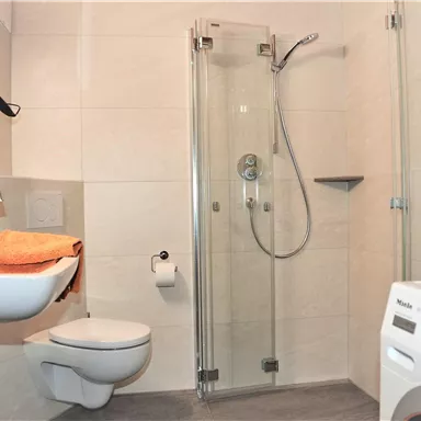 Ein modernes Badezimmer mit einer Duschkabine und einer Waschmaschine. Es gibt ein Waschbecken mit orangefarbenen Handtüchern und eine schlichte, helle Farbgestaltung.