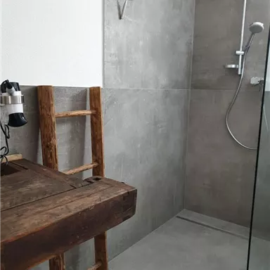 A modern bathroom with a glass shower cabin and gray tiles. In one corner, there is a wooden chair and a towel rack.