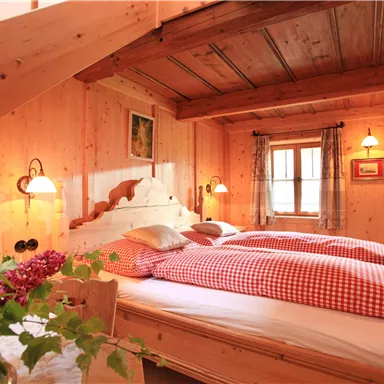 A cozy bedroom with wooden paneling and a large bed. The decor is rustic and inviting, with red checkered bed covers and a beautiful flower plant in the foreground.