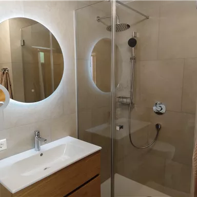 A modern bathroom with a glass shower and a wooden sink. A round mirror with lighting is mounted on the wall.