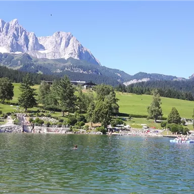 Ein schöner Badesee mit klarerem Wasser und Schwimmern. Im Hintergrund sind grüne Wiesen und majestätische Berge zu sehen.