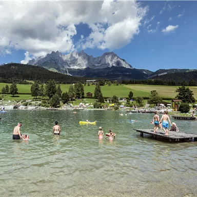 A clear lake surrounded by green meadows and mountains. People are swimming and relaxing by the water.