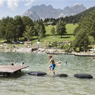 A boy is jumping on foam islands in the water. In the background, swimmers and a green meadow with mountains can be seen.