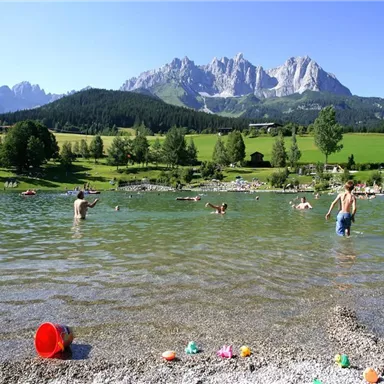 Ein schöner Badesee in den Bergen mit vielen Menschen, die im Wasser schwimmen. Im Hintergrund sind grüne Wiesen und beeindruckende Berggipfel zu sehen.