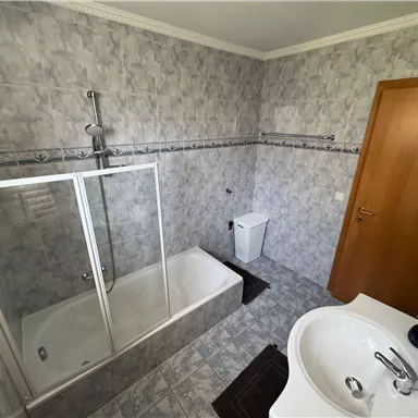 A modern bathroom with a bathtub and shower cabin. The walls are designed with gray tiles, and there is a sink as well as a toilet.
