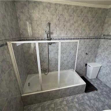A bathroom with a bathtub and shower cabin. The walls are tiled with gray tiles.