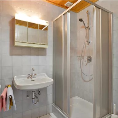 A modern bathroom with a shower, sink, and mirror. The walls are tiled in light colors and there are towels on a small shelf.