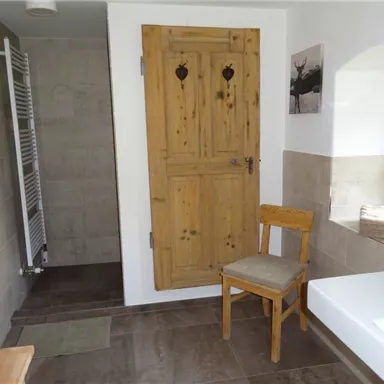 A modern bathroom with a wooden door and a chair. The walls are kept light, and in the background, a towel radiator is visible.