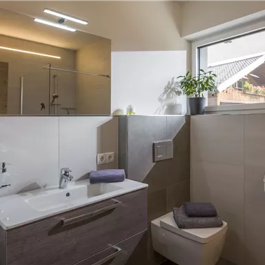 Ein modernes Badezimmer mit einem Waschbecken, einem Spiegel und einer Toilette. Die Fenster lassen natürliches Licht herein und es gibt eine kleine Pflanze auf der Fensterbank.