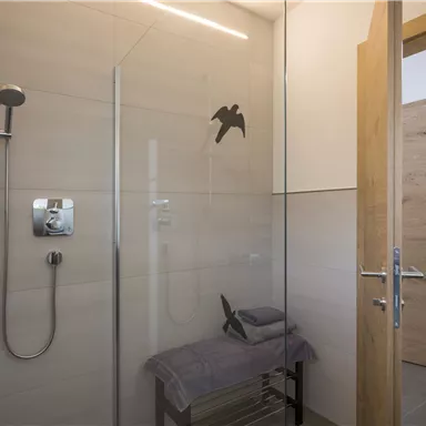 Ein modernes Badezimmer mit einer Glasdusche und eleganten Fliesen. Eine Bank mit Handtüchern ist im Raum platziert, und die Tür steht offen.