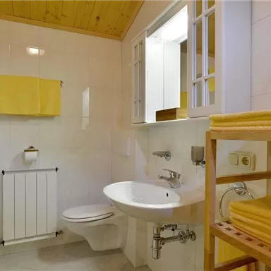 A modern bathroom with white walls and a wooden ceiling. Yellow towels are hanging for decoration, and there is a shower as well as a toilet.