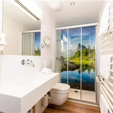 A modern bathroom with a sleek vanity and a toilet area. The shower wall features a picturesque landscape with mountains and a lake.
