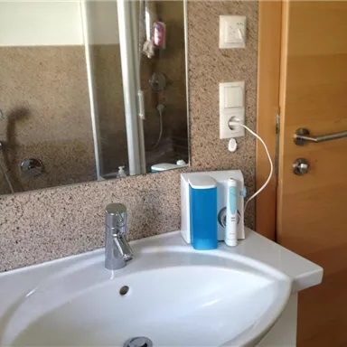 Ein Badezimmer mit einem Waschbecken und einem Spiegel. Im Hintergrund sind eine Dusche und eine Holzinnentür zu sehen.