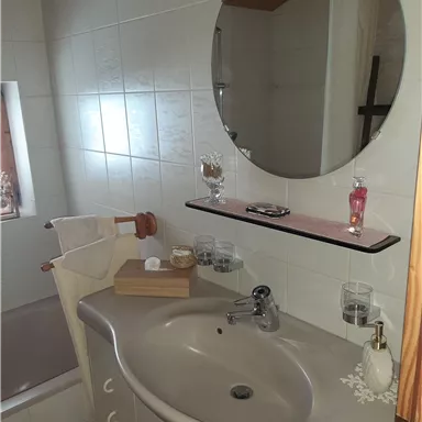A small bathroom with a round mirror above the sink. On the shelf are care products and a towel holder.