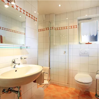 A bright bathroom with white tiles and a modern sink. There is a shower, a toilet, and a towel radiator.
